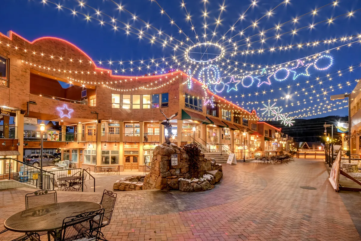 A lively outdoor plaza illuminated by string lights, featuring a stone fountain, inviting seating areas, and decorative storefronts. The setting hints at year-round socializing, shopping, dining, and community events in downtown Fraser.