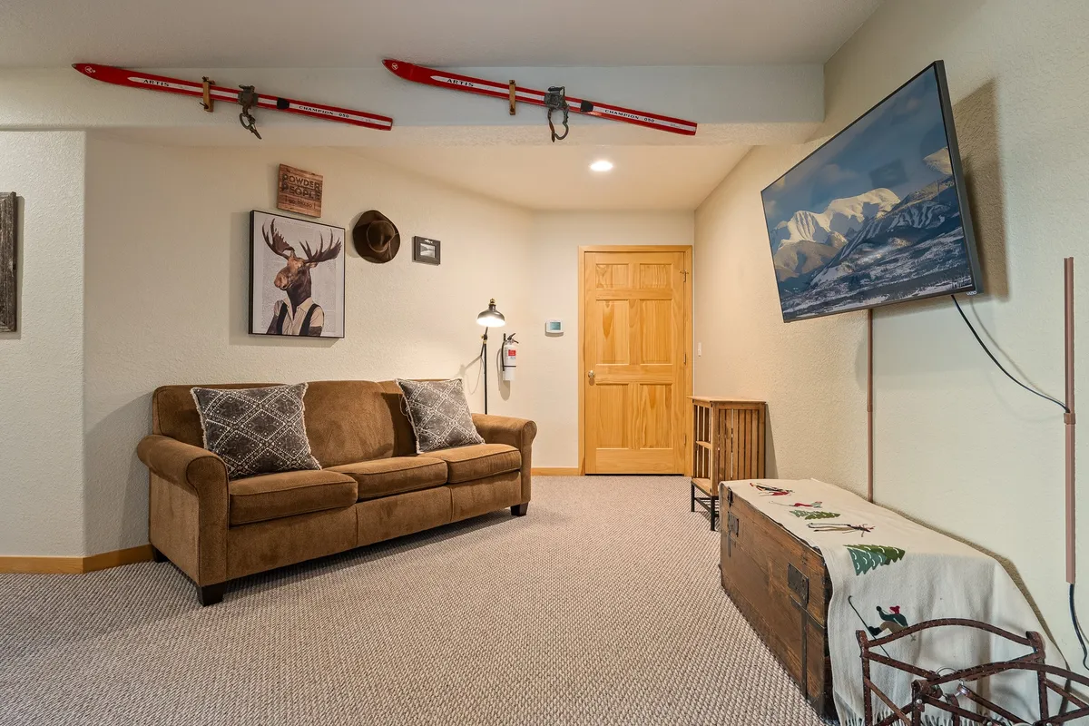 A spacious main living room featuring a comfortable sofa, ski-themed decoration, deer artwork, a large TV, and a wooden storage chest. The space is designed for relaxation, socializing, and entertainment, with a nod to local culture and outdoor adventure.