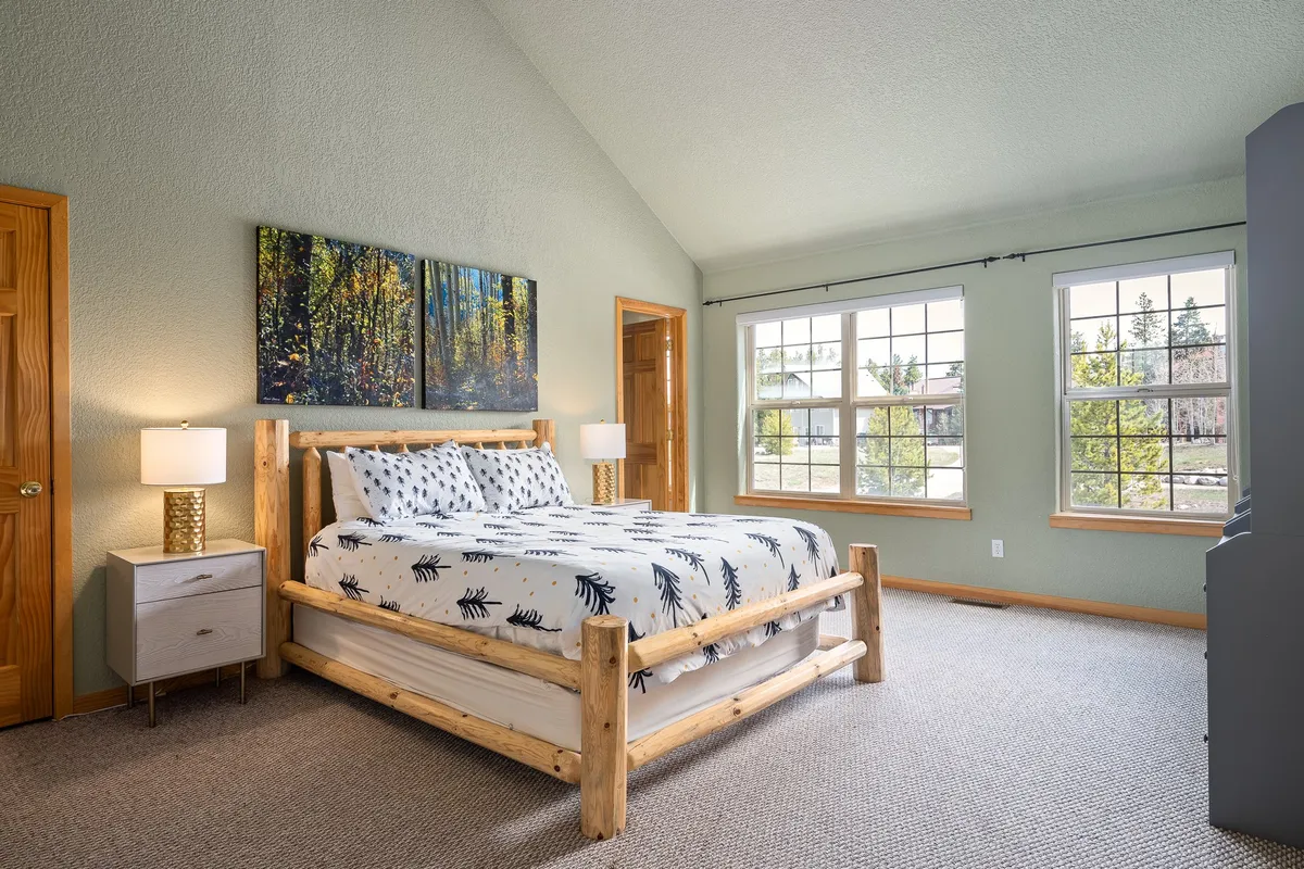 A comfortable and serene bedroom with a rustic wooden bed frame, forest-themed artwork, and large windows that fill the space with sunlight and views of surrounding trees.