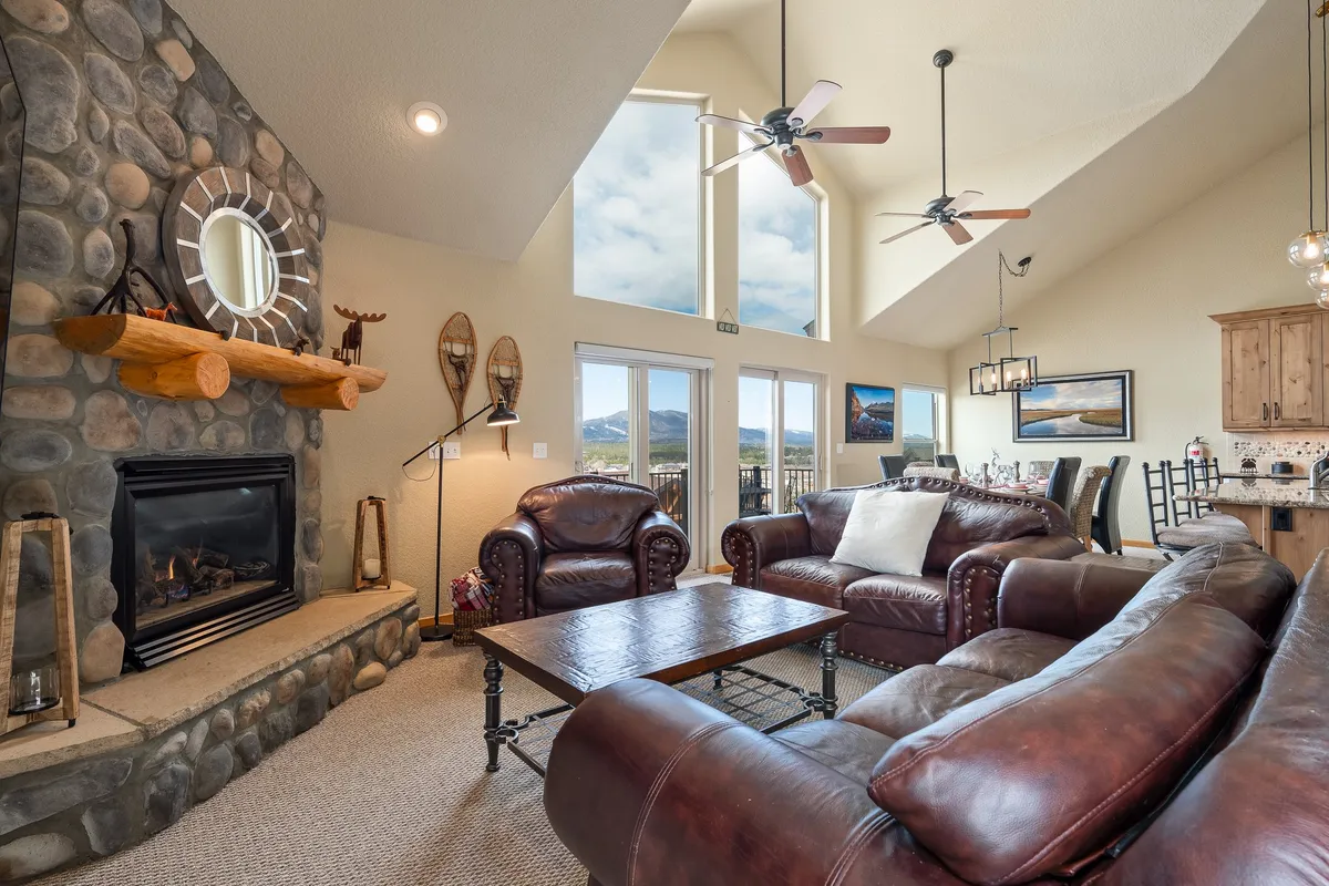 A beautiful open-concept living and dining area with a stone fireplace, leather sofas, and expansive windows that frame panoramic mountain views. The arrangement invites gatherings around the fire while also drawing the eye to the stunning scenery outside.