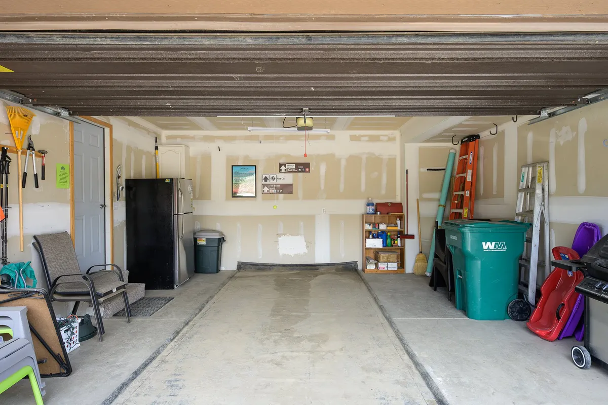 This image shows a practical and organized garage interior, complete with a refrigerator, green trash can, ladders, stacked chairs, and tools mounted on the wall. The space clearly functions as a multi-purpose area for vehicle parking, gear storage, and household organization.