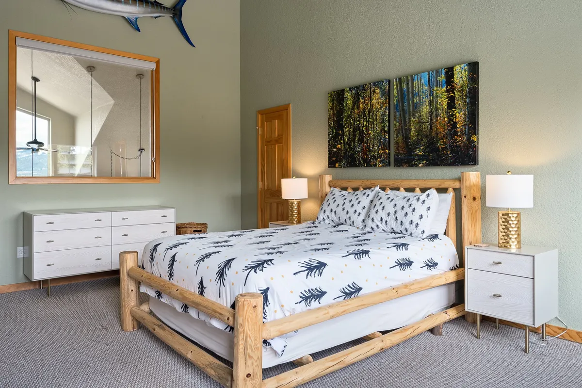 This image features a serene queen bedroom with a wooden frame bed, nature-themed artwork, mirrored wall, and nightstands with lamps. The room is bathed in natural light, creating a restful atmosphere ideal for sleep or quiet reading.
