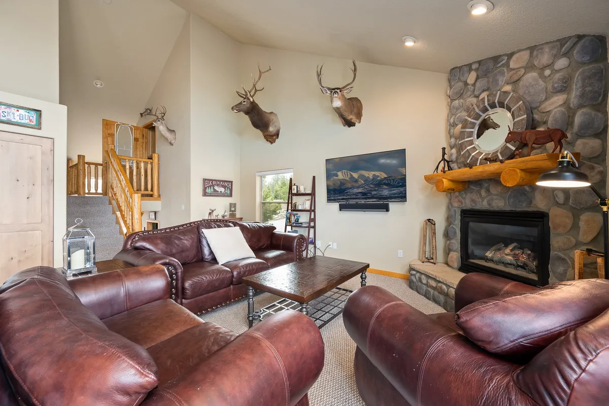 A warm, inviting lounge with a stone fireplace, mounted deer heads, leather furniture, and a wooden staircase. The space is perfect for group gatherings, relaxation, or watching TV.