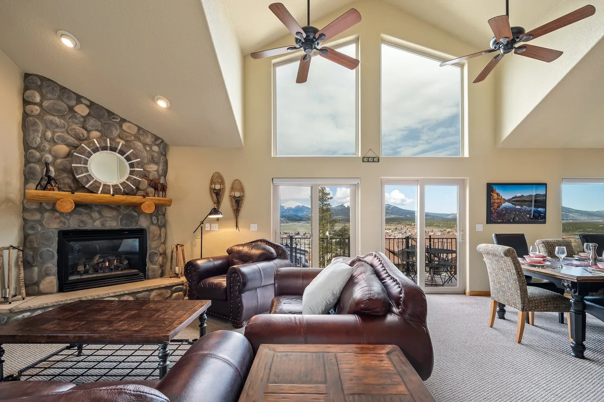 The image showcases a warm, inviting living space with a striking stone fireplace as the centerpiece, flanked by plush leather sofas and a spacious dining table. Large windows frame breathtaking sunset views, filling the room with natural light and creating a cozy, homey atmosphere perfect for gatherings or quiet evenings by the fire.