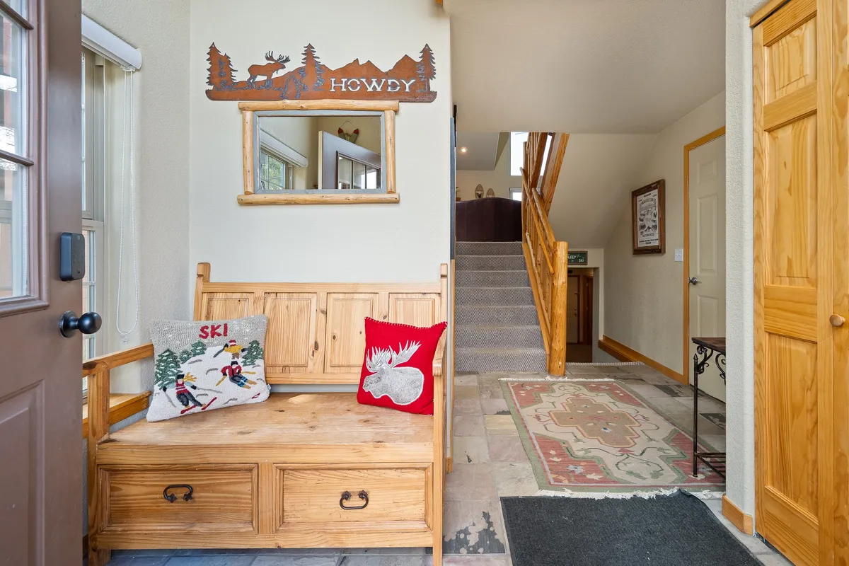 The image depicts a welcoming entryway with a rustic wooden bench adorned with decorative pillows, a mirror with a natural wood frame, and a staircase leading up. The space is bright, cozy, and inviting, setting the tone for the rest of the home.