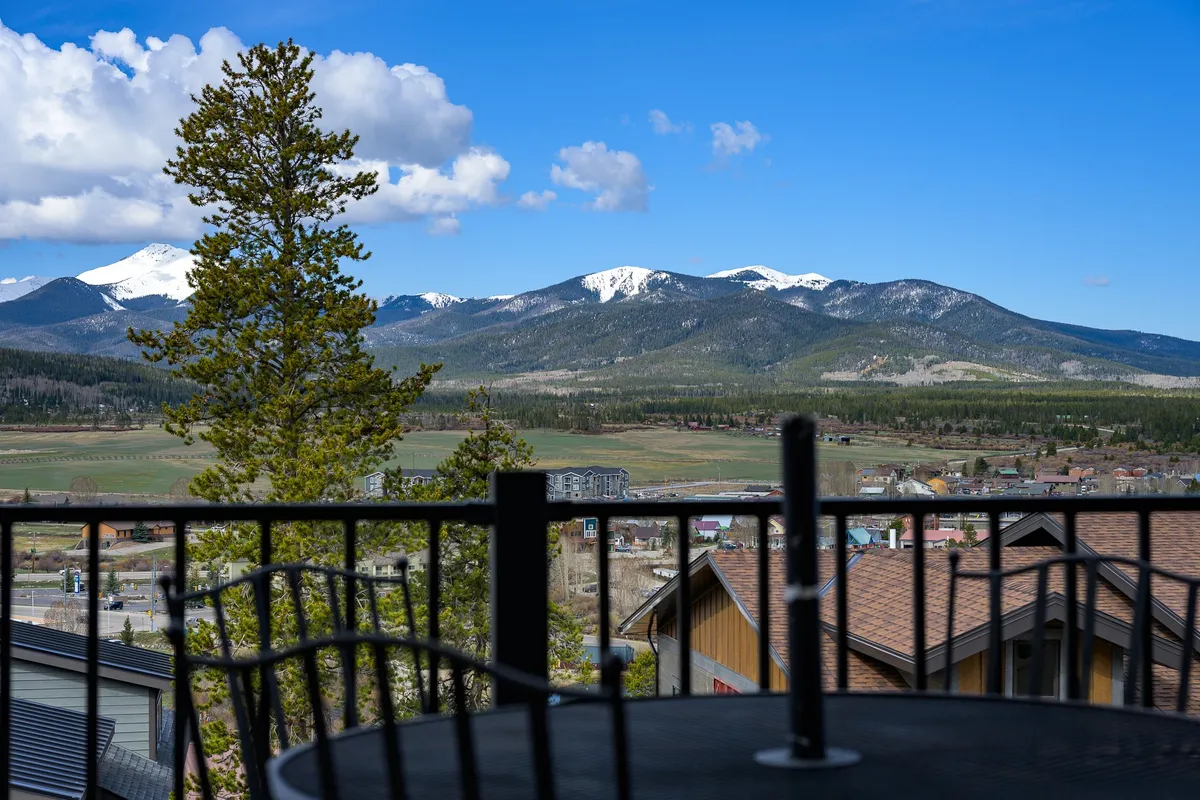 A panoramic view highlighting a majestic mountain range under a clear blue sky, with pine trees framing the scene and a charming town visible below. The vantage point suggests relaxation from a balcony or terrace, offering a perfect backdrop for leisure and unwinding after a day of adventure.