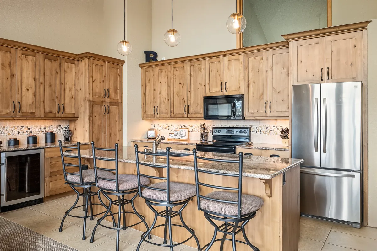 A spacious kitchen featuring a prominent island with bar stools, stainless steel appliances, and warm wood cabinetry. The island is set up for both food preparation and casual seating, while the appliances and layout highlight the space's modern functionality. The scene evokes a sense of social gathering and culinary activity.